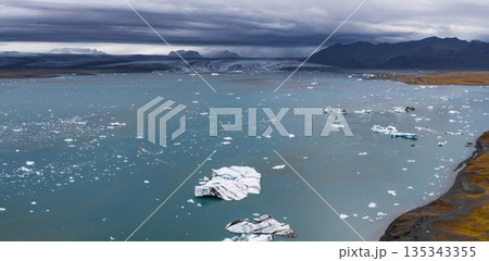 Turquoise waters of Jokulsarlon Glacier Lagoon with floating icebergs, a massive glacier, and dark mountains under a cloudy Icelandic sky. 135343355