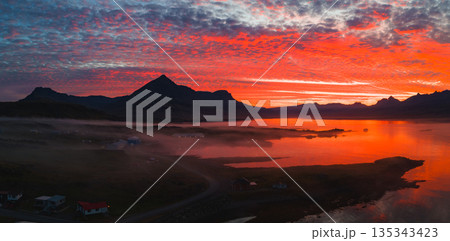 A vivid sunset illuminates an Icelandic scene with a mountain peak, calm water reflecting red and orange hues, scattered houses, and a winding road. A vivid sunset illuminates an Icelandic scene with a mountain peak, calm water reflecting red and orange hues, scattered houses, and a winding road. 135343423
