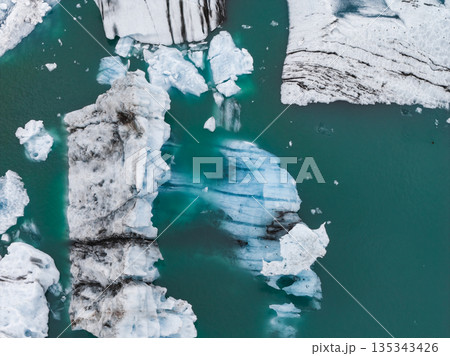 Large icebergs with black volcanic ash streaks and bright ice float in a turquoise glacial lagoon, showcasing dramatic contrasts and textures. Large icebergs with black volcanic ash streaks and bright ice float in a turquoise glacial lagoon, showcasing dramatic contrasts and textures. 135343426