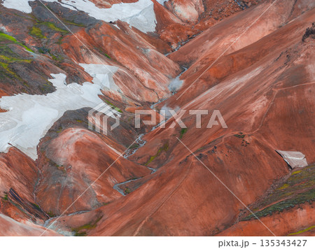 Vibrant red, orange, and brown rhyolite mountains with white snow patches and rising steam from geothermal vents in Iceland's highlands. Vibrant red, orange, and brown rhyolite mountains with white snow patches and rising steam from geothermal vents in Iceland's highlands. 135343427