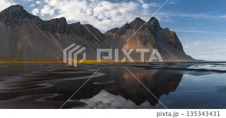 The Stokksnes Peninsula features the jagged Vestrahorn peaks mirrored in calm waters on a black sand beach under a partly cloudy blue sky. 135343431