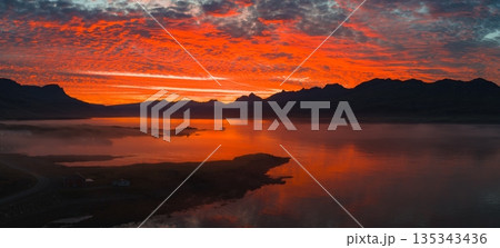 Vivid red and orange sunset reflects on still waters, with silhouetted peaks and mist. A cluster of buildings sits near the shoreline in the foreground. Vivid red and orange sunset reflects on still waters, with silhouetted peaks and mist. A cluster of buildings sits near the shoreline in the foreground. 135343436