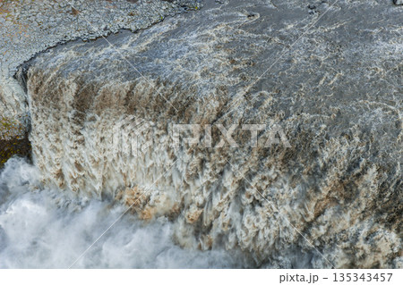 Dramatic aerial view of Dettifoss waterfall in Iceland, showcasing turbulent cascading water, white foam, brown sediment, and rugged rocky surroundings. Dramatic aerial view of Dettifoss waterfall in Iceland, showcasing turbulent cascading water, white foam, brown sediment, and rugged rocky surroundings. 135343457
