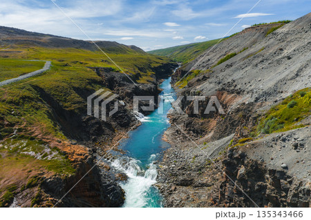 Turquoise river flows through Studlagil Canyon, flanked by basalt formations, green hills, and rocky terrain under a clear blue sky with scattered clouds. 135343466