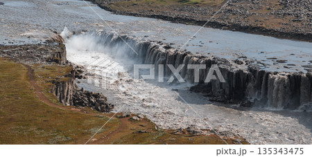 Dettifoss waterfall in Iceland cascades over rugged cliffs, surrounded by basalt formations, mist, and a rocky landscape with a visible dirt path. 135343475