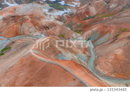 Vibrant rhyolite hills in Landmannalaugar, Iceland, with orange, red, and brown hues, green moss, snow patches, a winding trail, and a small stream. 135343485
