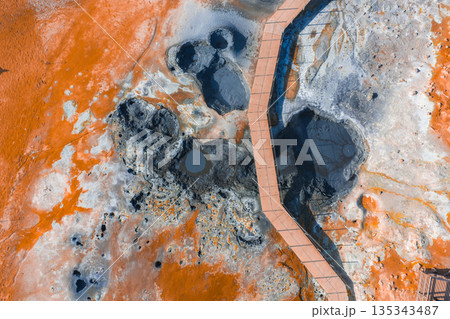 Aerial image of a geothermal area in Iceland featuring orange and white mineral deposits, steaming fumaroles, bubbling mud pots, and a wooden boardwalk. Aerial image of a geothermal area in Iceland featuring orange and white mineral deposits, steaming fumaroles, bubbling mud pots, and a wooden boardwalk. 135343487