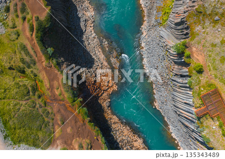 Studlagil Canyon in Iceland features a turquoise river, dramatic basalt columns, lush greenery, and a viewing platform on the right side. Studlagil Canyon in Iceland features a turquoise river, dramatic basalt columns, lush greenery, and a viewing platform on the right side. 135343489