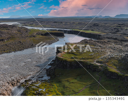 Dettifoss waterfall cascades over rugged cliffs in Iceland, surrounded by rocky terrain, green vegetation, and a winding path under a partly cloudy sky. 135343491