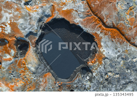 Geothermal landscape in Iceland featuring a dark pool, vibrant orange and rust mineral deposits, visible cracks, and textured earth patterns. 135343495