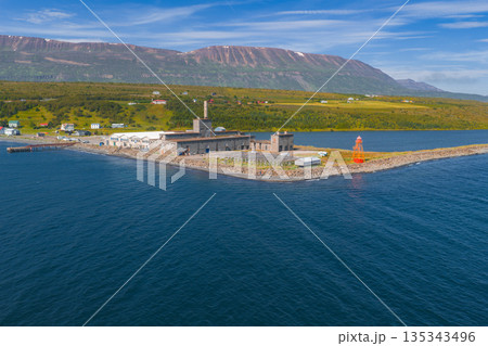 Aerial view of an Icelandic coastal area with a tall chimney, orange lighthouse, green fields, rugged mountains, calm blue water, and clear sky. 135343496