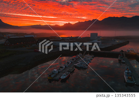 A small harbor in Iceland at sunset, featuring boats, industrial buildings, traditional houses, jagged mountains, and a misty, vibrant sky. 135343555