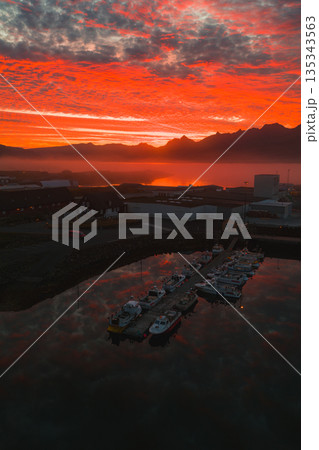 Aerial view of a small harbor in Iceland during sunset. Boats dock along a pier as fiery red and orange hues reflect on calm water with mountain silhouettes. 135343563