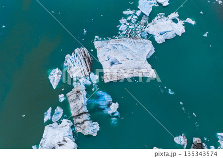 Large icebergs with white, blue, and black patterns float in a turquoise glacial lagoon, showcasing unique shapes and glacial sediment in Iceland. Large icebergs with white, blue, and black patterns float in a turquoise glacial lagoon, showcasing unique shapes and glacial sediment in Iceland. 135343574