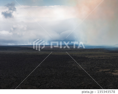 Dark volcanic terrain with cooled lava fields stretches into the distance under low clouds and mist, with hints of orange and blue in the sky. 135343578
