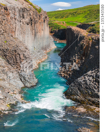 Studlagil Canyon features dramatic basalt columns, a turquoise river, and lush green surroundings, showcasing Iceland's volcanic landscape. 135343580