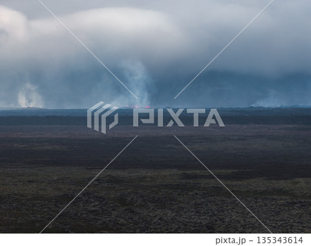 Volcanic eruption in Iceland with glowing red lava, thick smoke, and a rocky foreground. Dense clouds dominate the background, creating a dramatic scene. Volcanic eruption in Iceland with glowing red lava, thick smoke, and a rocky foreground. Dense clouds dominate the background, creating a dramatic scene. 135343614