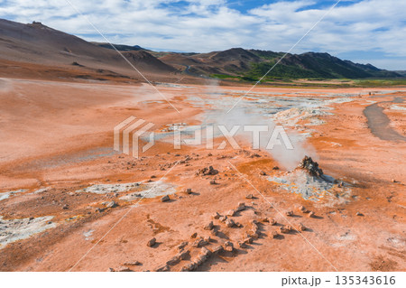 A geothermal landscape in Iceland featuring orange and red earth tones, steam from fumaroles, rocky terrain, and green hills under a blue sky. 135343616