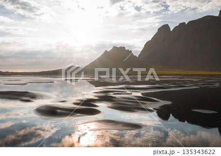 The Stokksnes Peninsula features the Vestrahorn mountains, a black sand beach, and shallow water pools reflecting the sky under soft sunlight. 135343622