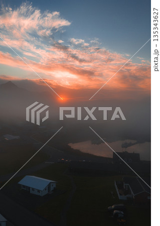 A serene sunset illuminates a misty Icelandic scene with a white roofed house, winding roads, grassy terrain, calm water, and fog covered mountains. 135343627