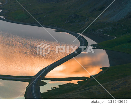 A serene aerial view of a winding road dividing a reflective lagoon during sunset, with orange skies mirrored in the water and rocky green terrain. 135343633
