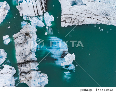 Icebergs with black volcanic ash and blue ice float in a turquoise glacial lagoon. The fractured shapes and vibrant water highlight Iceland's geology. 135343638