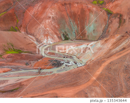 Aerial view of Iceland's Rainbow Mountains featuring vibrant red, orange, and green hues, a winding stream, and a small wooden bridge crossing it. 135343644