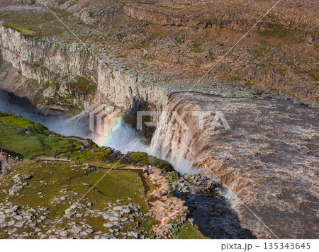 Dettifoss waterfall cascades powerfully, creating mist and a vivid rainbow. Rugged cliffs, green vegetation, and a viewing path with visitors are visible. 135343645