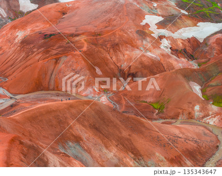 Vibrant multicolored rhyolite hills with red, orange, and brown hues, green moss, snow patches, winding trails, and small figures in the vast landscape. Vibrant multicolored rhyolite hills with red, orange, and brown hues, green moss, snow patches, winding trails, and small figures in the vast landscape. 135343647