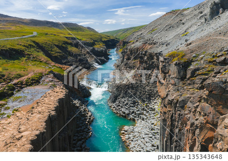 Studlagil Canyon in Iceland features dramatic basalt columns, a turquoise river, earthy canyon walls, green hills, and a winding road under a clear sky. 135343648