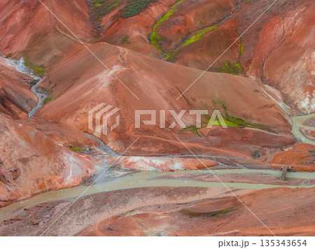 Multicolored rhyolite hills of Iceland's Rainbow Mountains with red, orange, and green hues. A wooden bridge crosses a river, with steam rising nearby. 135343654