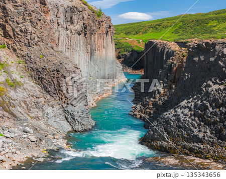 Studlagil Canyon in Iceland features towering basalt columns, a vibrant turquoise river, lush green surroundings, and a clear blue sky with clouds. Studlagil Canyon in Iceland features towering basalt columns, a vibrant turquoise river, lush green surroundings, and a clear blue sky with clouds. 135343656