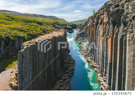 Studlagil Canyon in Iceland features dramatic basalt columns, a vibrant turquoise river, lush green landscapes, and rolling hills under a blue sky. 135343675