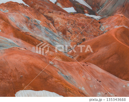 Vibrant orange and red rhyolite hills with snow patches and steaming vents in Iceland's Kerlingarfjoll, with hikers visible on rugged ridges. 135343688