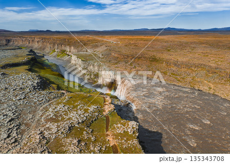 Aerial view of Dettifoss waterfall in Iceland, with mist creating a faint rainbow. Rugged cliffs and barren highlands surround the powerful cascade. 135343708