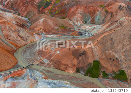 Vibrant rhyolite mountains in Landmannalaugar, Iceland, with a winding river, wooden bridge, moss patches, and hikers on a scenic trail. 135343719