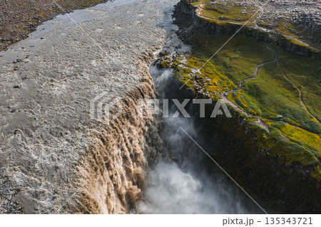 Dettifoss waterfall cascades into a deep canyon with misty spray, surrounded by moss, grass, rocky terrain, and a winding path to a viewpoint. 135343721