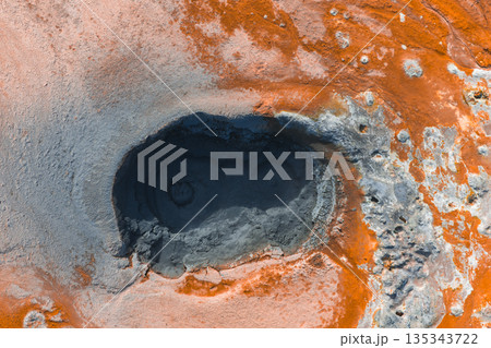 Aerial view of a dark mud pool surrounded by orange and reddish brown terrain, showcasing geothermal activity and textured mineral deposits in Iceland. Aerial view of a dark mud pool surrounded by orange and reddish brown terrain, showcasing geothermal activity and textured mineral deposits in Iceland. 135343722