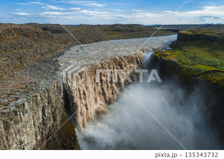 Dettifoss waterfall cascades into a deep canyon, with mist rising prominently. Rugged rocks, green moss, and small figures highlight its scale. 135343732