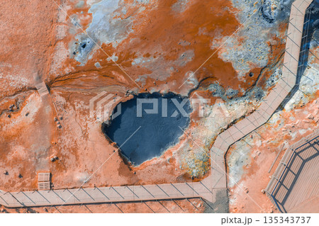Geothermal area in Iceland with vibrant orange and red earth tones, a dark bubbling mud pool, and wooden boardwalks winding through the landscape. 135343737