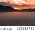 A vibrant sunset illuminates the sky with orange and red hues, silhouetting rugged mountains. Dense mist blankets the foreground, creating a serene scene. 135343739