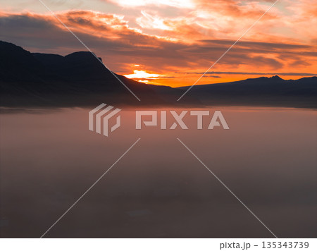 A vibrant sunset illuminates the sky with orange and red hues, silhouetting rugged mountains. Dense mist blankets the foreground, creating a serene scene. A vibrant sunset illuminates the sky with orange and red hues, silhouetting rugged mountains. Dense mist blankets the foreground, creating a serene scene. 135343739