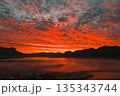 Fiery red and orange sunset over a calm body of water in Iceland, with silhouetted mountains, textured clouds, and a misty settlement in the foreground. 135343744