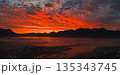 Fiery red and orange sunset over Icelandic mountains, mist covered ground, and a calm reflective body of water with a small settlement visible. 135343745