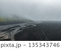 Aerial view of a volcanic landscape in Iceland with dark terrain, winding glacial rivers, and a moss covered mountain emerging from dense fog. 135343746