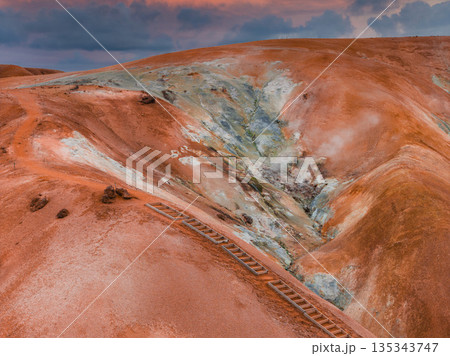 Vivid orange and red hills with white and green mineral deposits, a wooden staircase, and a dramatic pink and purple sky in Iceland's geothermal area. 135343747