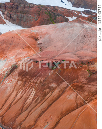 Aerial view of Iceland's Rainbow Mountains, featuring vibrant red, orange, and brown rhyolite formations with contrasting snow patches in the background. 135343752