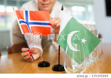 Young woman putting flags of Norway and Pakistan on table in office Young woman putting flags of Norway and Pakistan on table in office 135345894
