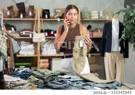 Girl is talking on phone while choose trousers in store department Girl is talking on phone while choose trousers in store department 135345943