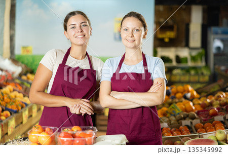 Happy two female sellers wearing apron and standing next to organic foods in supermarket Happy two female sellers wearing apron and standing next to organic foods in supermarket 135345992